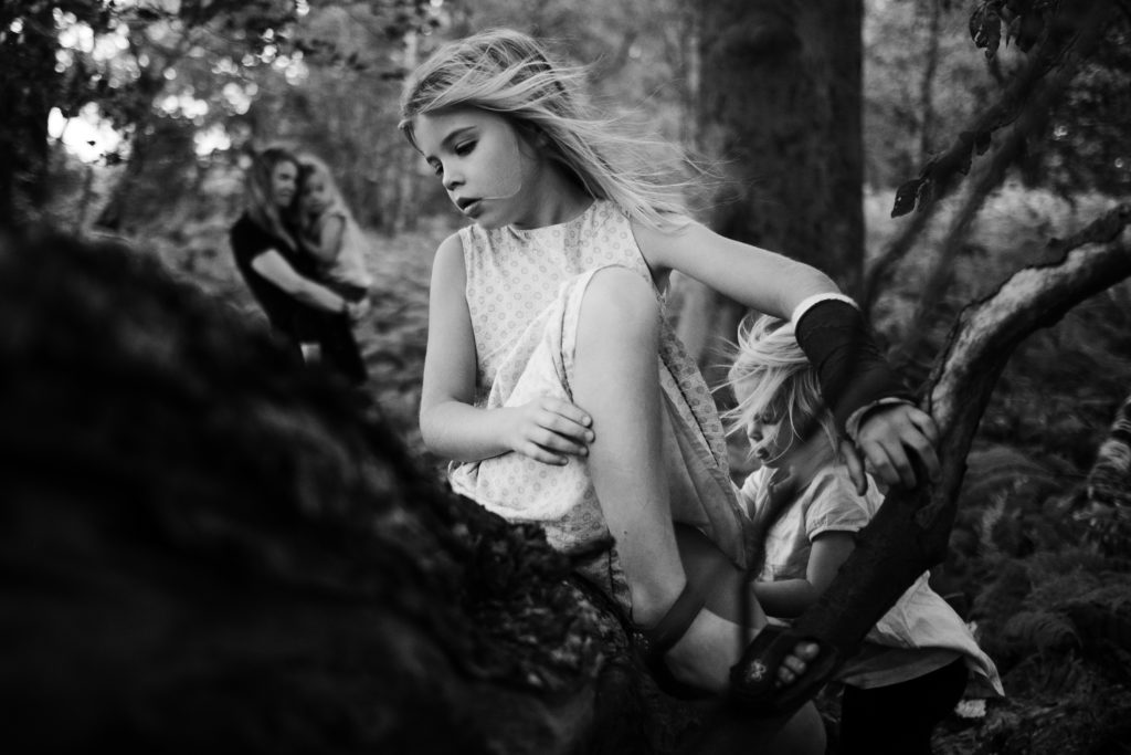 im Hintergrund steht eine Mutter mit ihrer Tochter auf dem Arm, im Vordergrund ist ein Baumstamm zu sehen, auf dem Baumstamm sitzt ein Mädchen, neben ihr steht ihre kleine Schwester, der Wnd weht ihr Haar aus dem Gesicht, sie sind im Wald