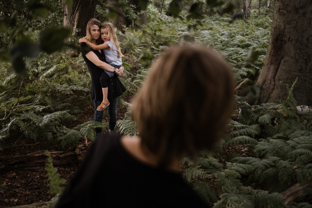 eine Mutter hat ihre Tochter auf dem Arm, sie stehem im Wald, im Vordergrund ist der Rücken eines Jungen zu sehen