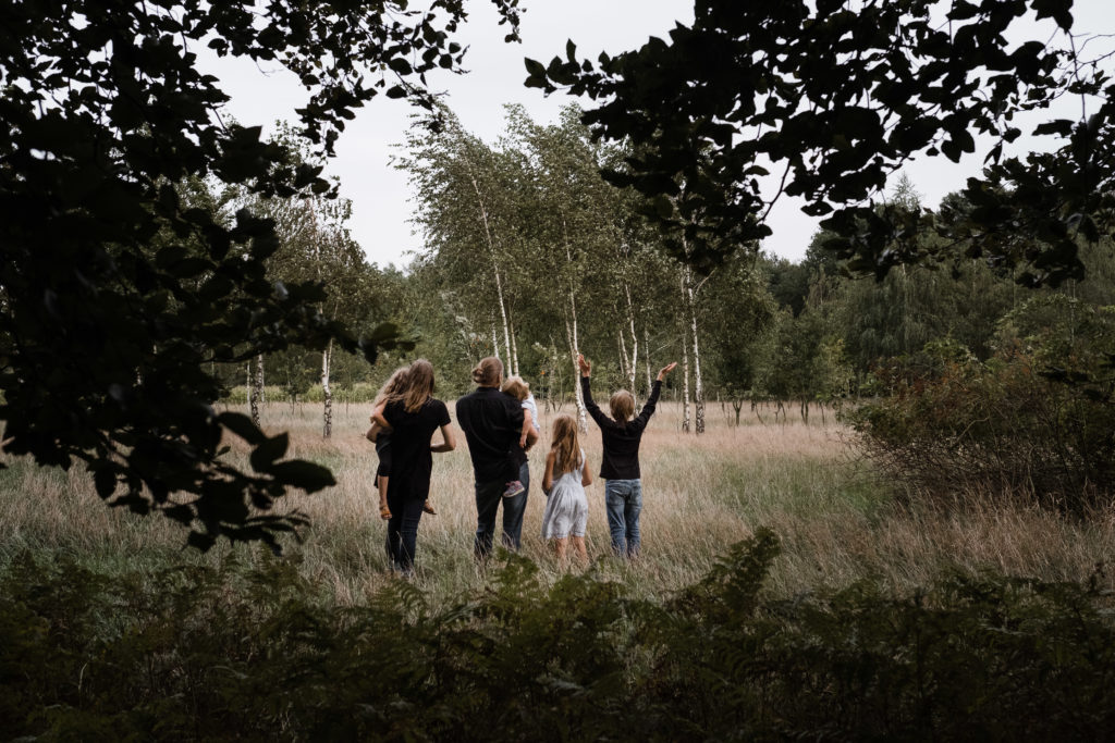auf einer Wiese, die umgeben von Wald ist, stehen eine Mutter und ein Vater mit ihren 3 Töchtern und ihrem Sohn, sie schauen in die Wiese, der Sohn hebt die Hände