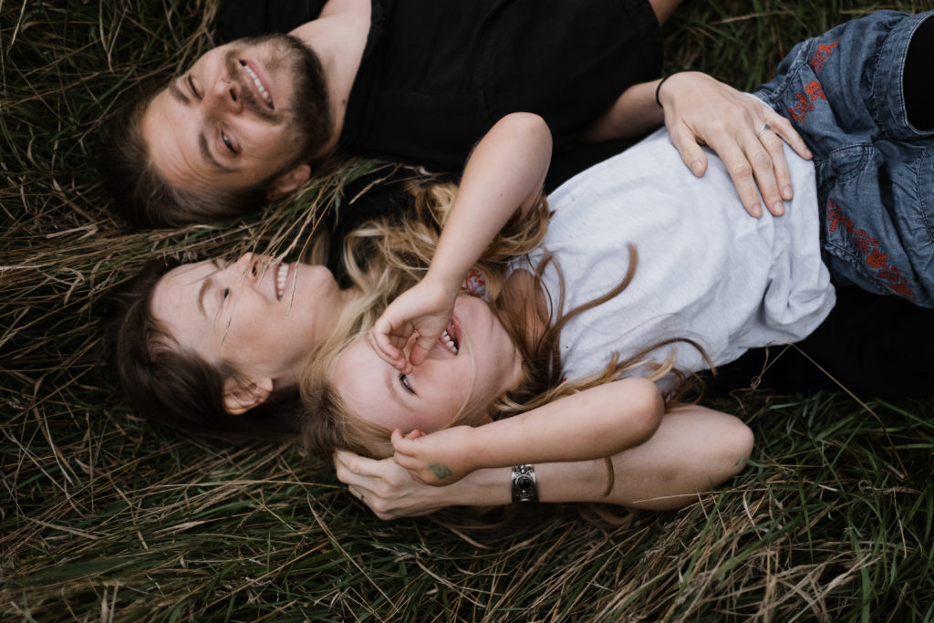 auf einer Wiese liegen eine Mutter und ein Vater mit ihrer Tochter, sie lachen