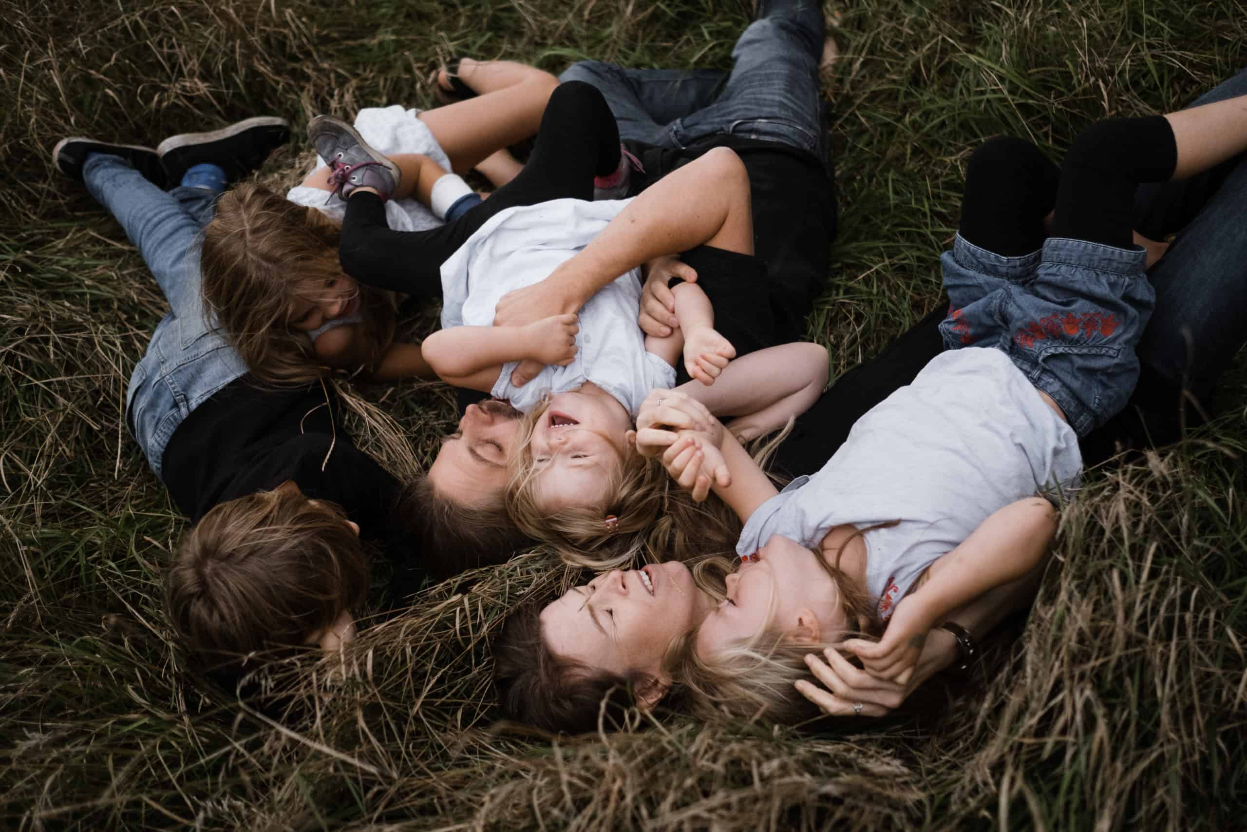 eine Mutter und ein Vater liegen mit ihren 3 Töchtern und ihrem Sohn auf einer Wiese, sie liegen kreuz und quer und auch übereinander, sie lachen