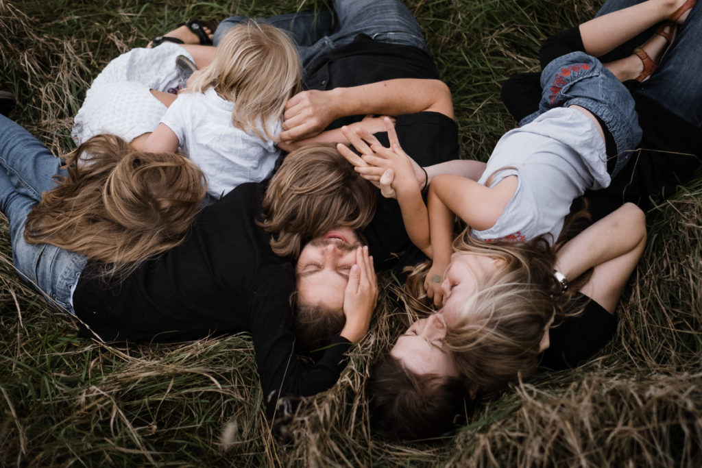 eine Mutter und ein Vater liegen mit ihren 3 Töchtern und ihrem Sohn auf einer Wiese, sie liegen kreuz und quer und auch übereinander, sie lachen