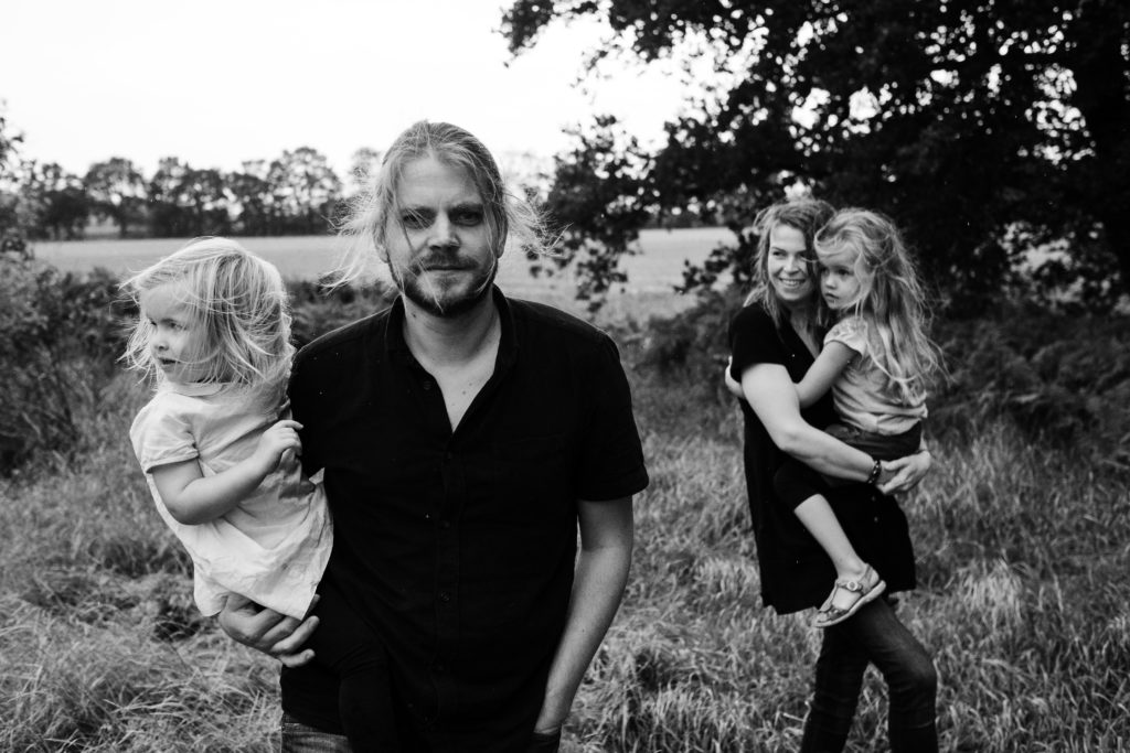 ein Vater hat seine Tochter auf dem Arm, hinter ihm geht seine Frau, die auch eine Tochter auf dem Arm trägt, sie gehen entlang eines Waldrandes