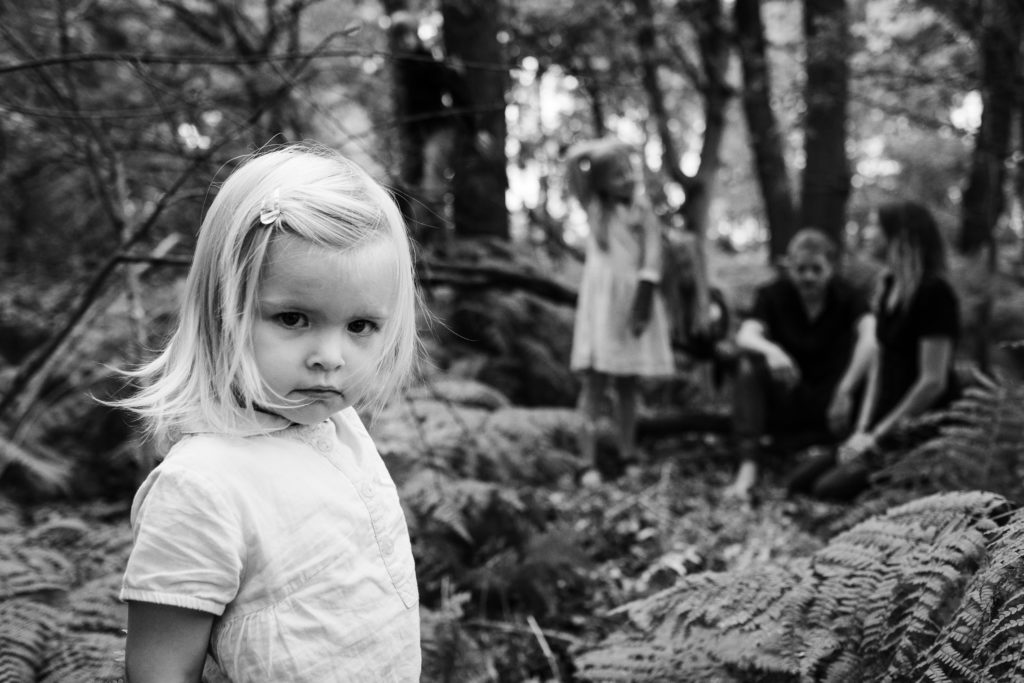 im Wald steht im Vordergrund ein kleines Mädchen, hinter ihr steht ihre Schwester, sowie ihre Mutter und ihr Vater, der Boden ist voller Fahn