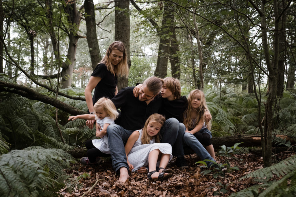 im Wald sitzen eine Mutter und ein Vater mit ihren 3 Töchtern und ihrem Sohn auf einem Baumstamm