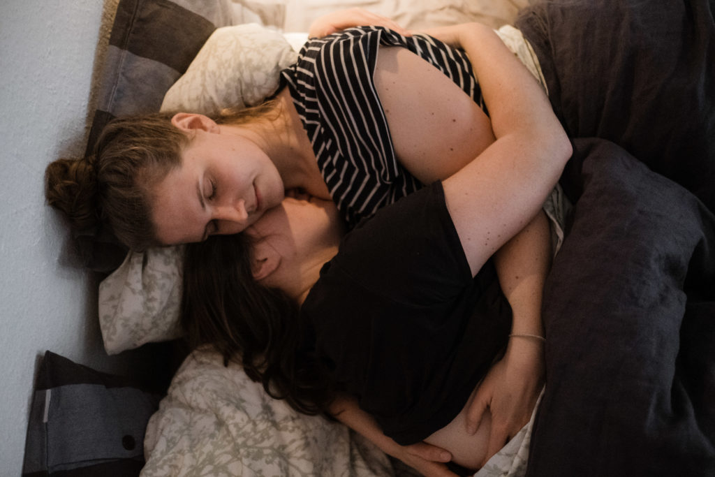 2 Frauen liegen auf einem Bett, sie halten sich in den Armen, eine Frau legt ihren Kopf in den Hals der anderen Frau, ihre Augen sind geschlossen