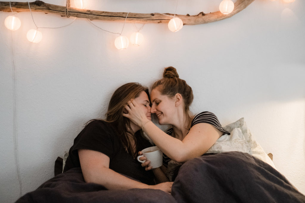 2 Frauen sitzen in einem Bett, über ihnen hängt eine Lampion- Lichterkette, eine Frau berührt die andere Frau im Gesicht, in der anderen Hand hält sie eine Tasse, ihre Stirn halten sie aneinander