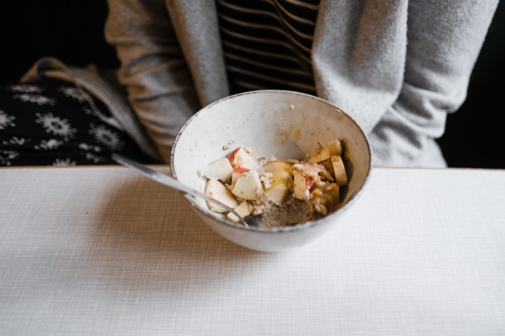 an einem weißen Tisch sitzt eine Frau, ihre Hände liegen auf ihrem Schoß, vor ihr steht eine Schüssel, die mit Äpfeln und Müsli gefüllt sind
