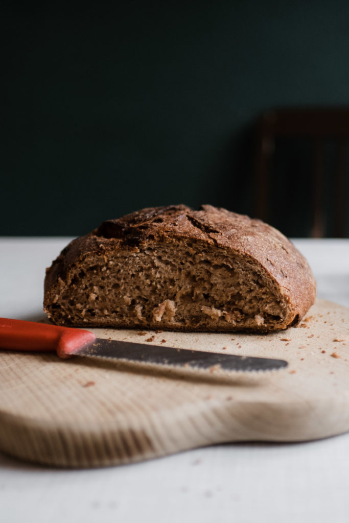 auf einem Tisch liet ein Holzbrett, auf dem ein Laib Brot liegt , davor liegt ein Messer und Brotkrümel