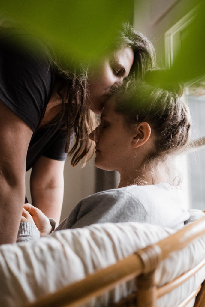 eine Frau sitzt auf einem Rattansofa, sie schließt die Augen, eine andere Frau lehnt vor ihr und küsst sie auf die Stirn