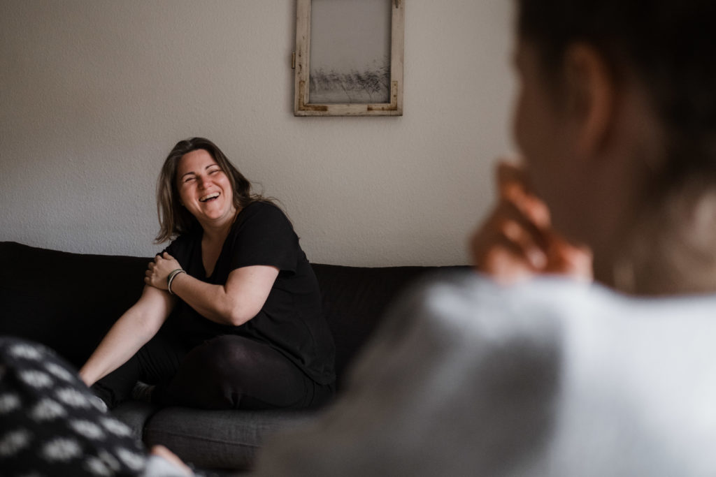 vor einer hellen Wand sitzt eine Frau auf einem Sof, sie lacht und hält sich dabei den Arm, ihr gegenüber sitzt eine andere Frau, ihr Blick richtet sich zu ihr