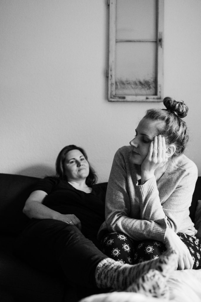 2 Frauen sitzen auf einem Sofa, eine Frau stützt ihre Beine auf einem Sessel ab, die andere Frau sitzt mit aufrechtem Oberkörpr auf dem Sofa, sie stützt ihren Kopf an ihrer Hand ab