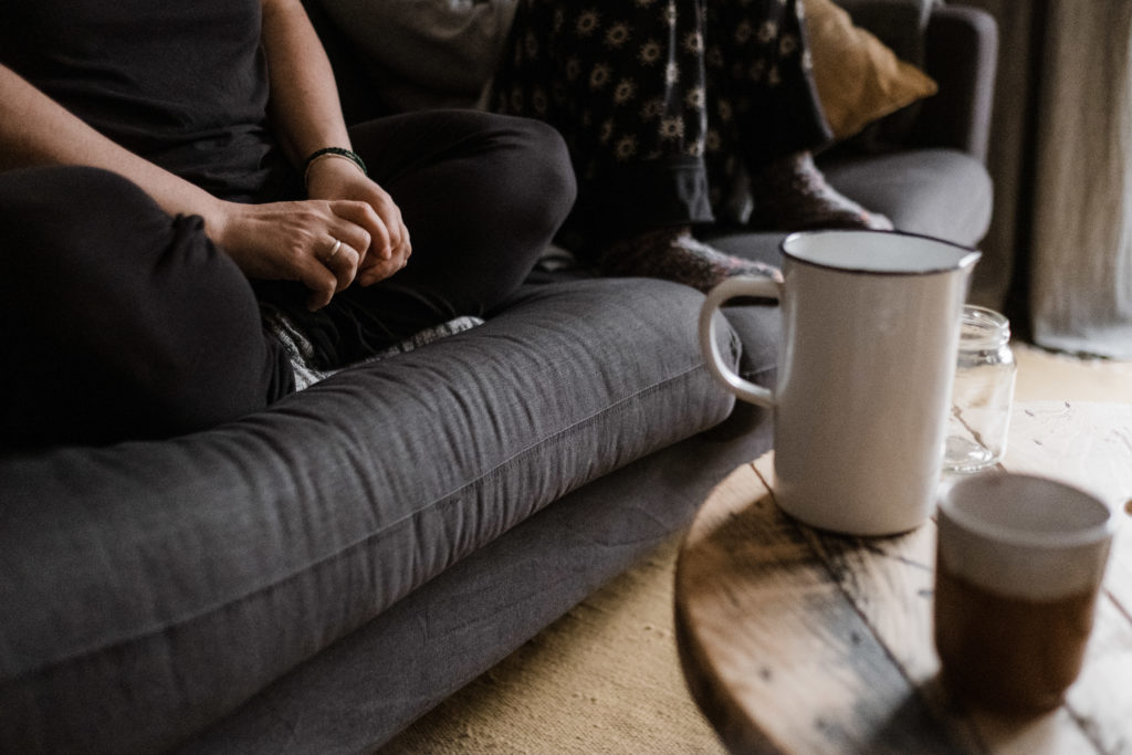 2 Frauen sitzen auf einem Sofa, eine Frau sitzt im Schneidersitz, die andere Frau sitzt mit aufgestellten Beinen auf dem Sofa