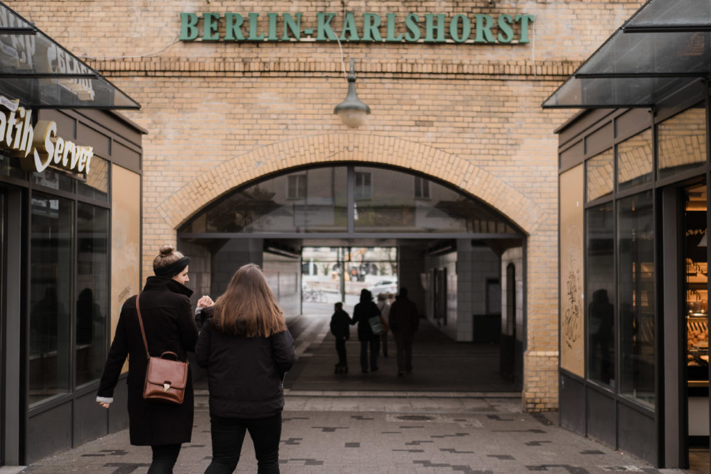 2 Frauen gehen in Richtung eines Gebäude ( Berlin- Karlshorst) , das Gebäude hat einen großen Glasbogen, die beiden Frauen halten sich an den Händen