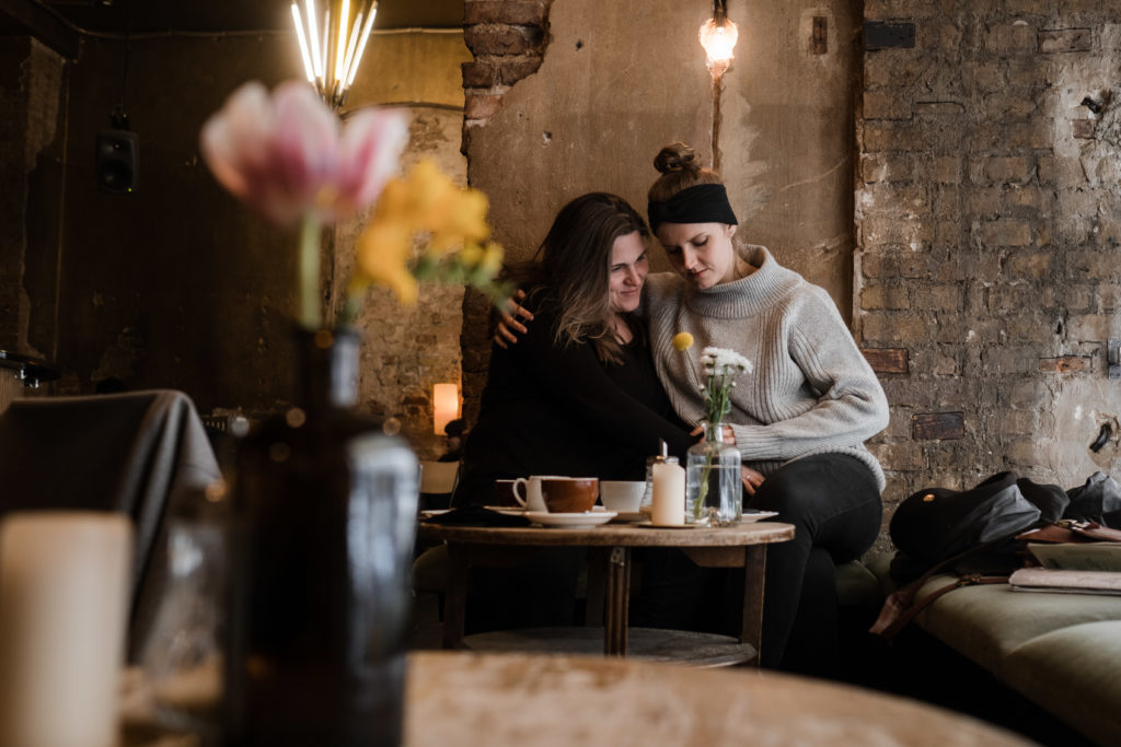 2 Frauen sitzen in einem Gebäude vor einer Steinwand, eine Frau legt ihren Arm und die andere Frau, vor ihnen steht eine Holzkabeltommel, auf der Tassen und eine Blumenvase stehen