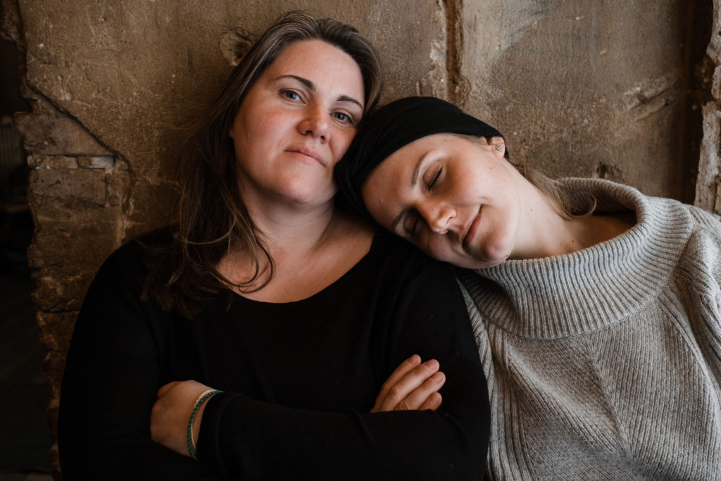 2 Fraun sitzen vor einer Steinwand in einem Gebäude, eine Frau verschränlt die Arme, die andere Frau lehnt ihren Kopf auf die Schulter der Frau