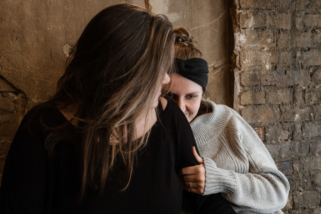 2 Fruen sitzen vor einer Steinwand in einem Gebäude, ein Frau hält ihren Kopf an den Rücken der anderen Frau, die andere Frau schaut zu ihr nach hinten