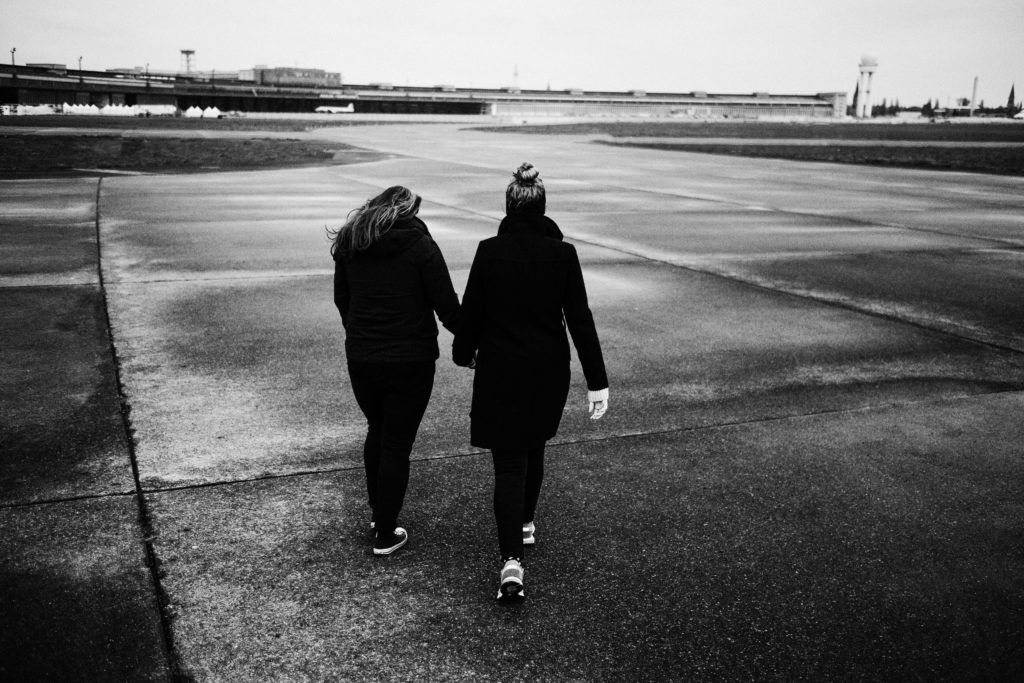 auf dem Rollfeld des alten Flughafen in Berlin laufen 2 Frauen, beide in schwarz gekleidet, sie halten gegenseitig ihre Hände, man sieht sie von hinten