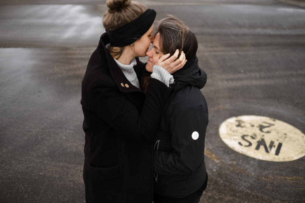 2 Frauen stehen auf dem Rollfeld des alten Flughafen in Berlin, sie sind beide schwarz gekleidet, sie halten sich in den Armen, eine Frau hält ihre Hand an das Ohr der anderen Frau, ihre Gesichter berühren sich