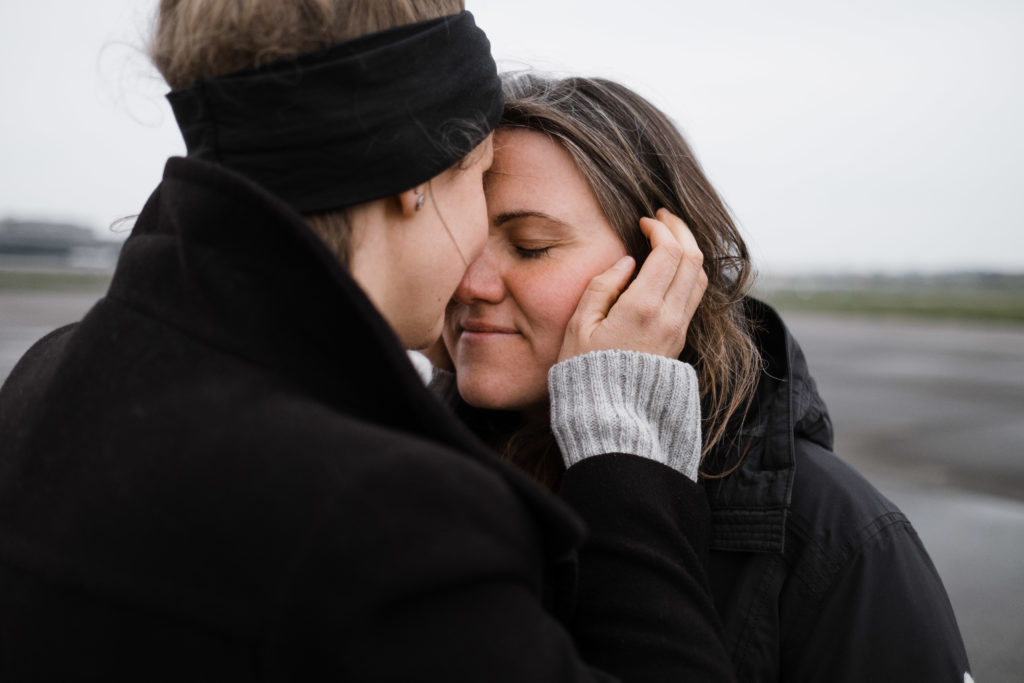 2 Frauen stehen auf dem Rollfeld des alten Flughafen in Berlin, sie sind beide schwarz gekleidet, sie halten sich in den Armen, eine Frau hält ihre Hand an das Ohr der anderen Frau, ihre Gesichter berühren sich, beide schließen die Augen
