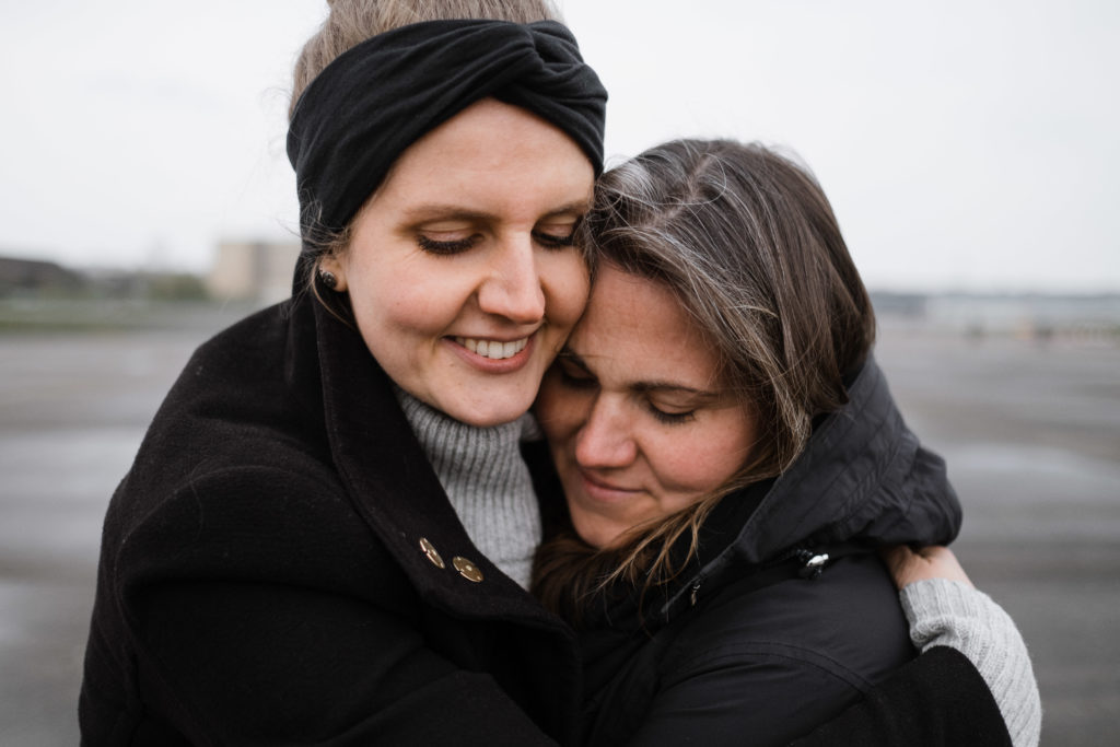 2 Frauen stehen auf dem Rollfeld des alten Flughafen in Berlin, sie sind beide schwarz gekleidet, sie umarmen sich und halten ihre Köpfe nebeneinander, beide schließen die Augen