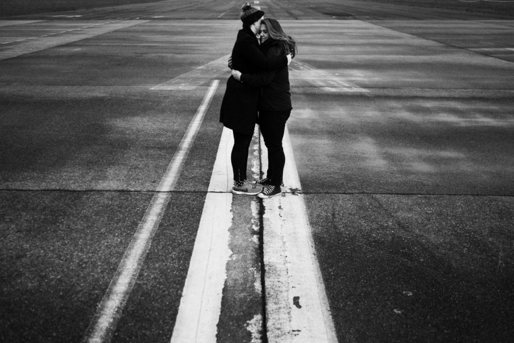 2 Frauen stehen auf dem Rollfeld des alten Flughafen in Berlin, sie sind beide schwarz gekleidet, sie stehen auf einem großen weißen Streifen, der auf dem Boden ist, sie umarmen sich