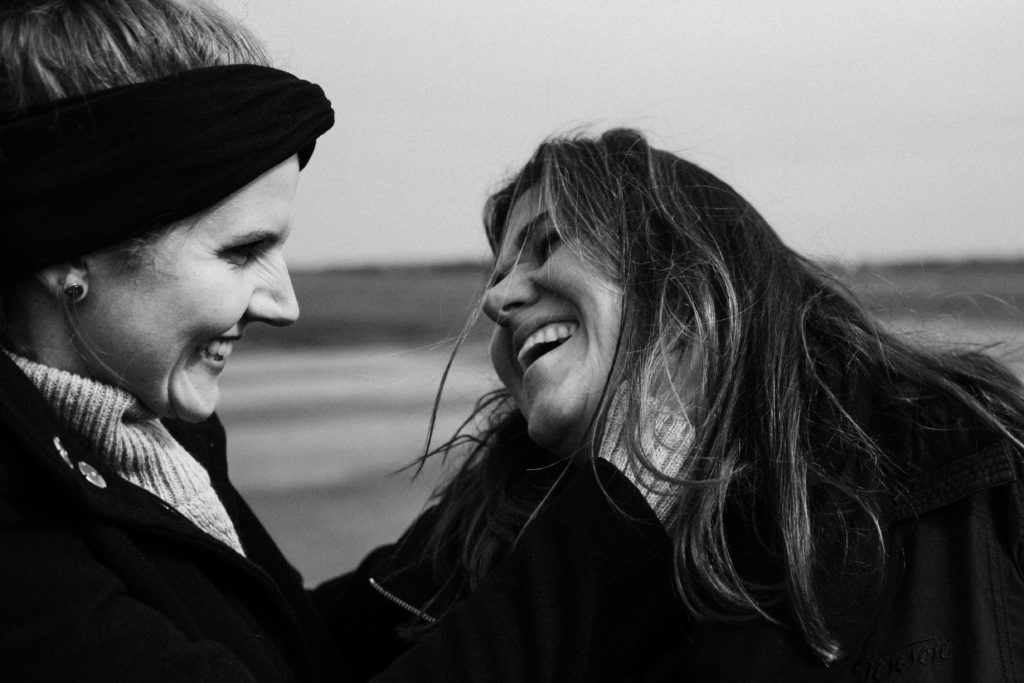 2 Frauen stehen auf dem Rollfeld des alten Flughafen in Berlin, sie sind beide schwarz gekleidet, eine Frau hält ihre Hand an das Ohr der anderen Frau, beide lachen