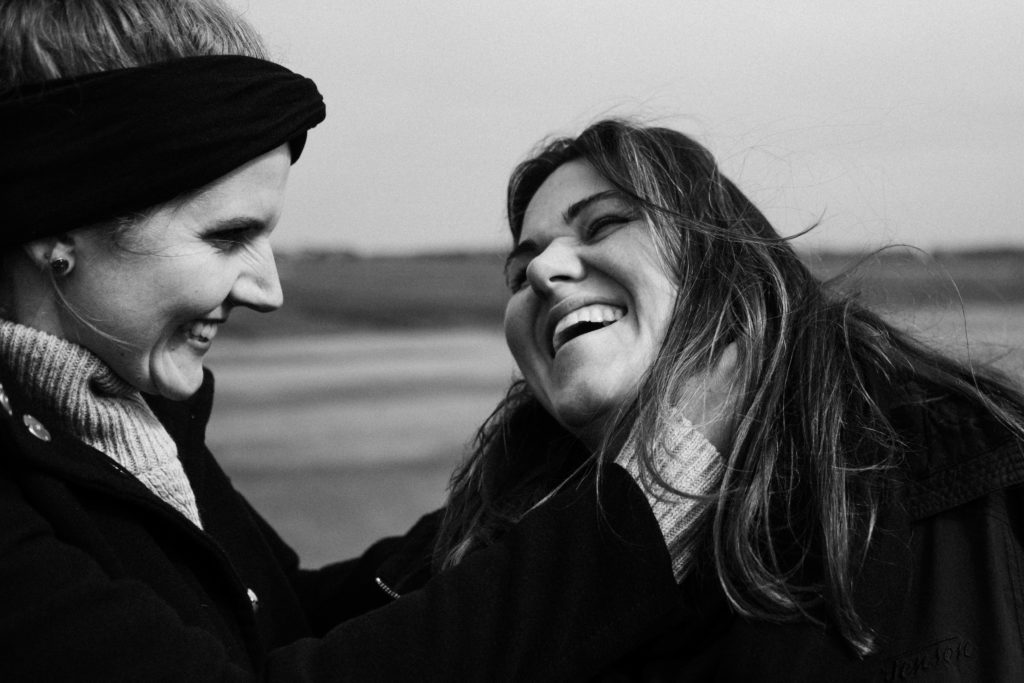 2 Frauen stehen auf dem Rollfeld des alten Flughafen in Berlin, sie sind beide schwarz gekleidet, eine Frau hält ihre Hand an das Ohr der anderen Frau, beide lachen