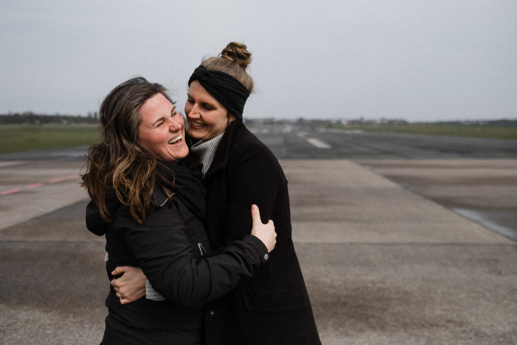 2 Frauen stehen auf dem Rollfeld des alten Flughafen in Berlin, sie sind beide schwarz gekleidet, sie umarmen sich, beide lachen