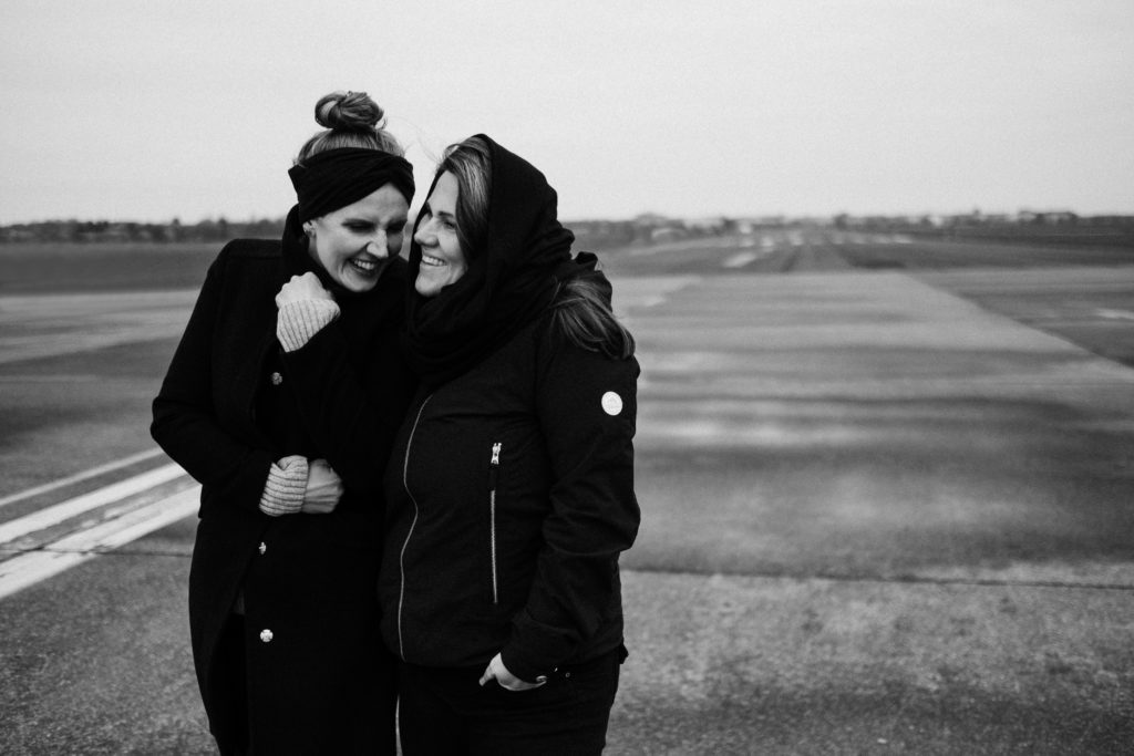 auf dem alten Rollfeld des Flughafen in Berlin steh2 Frauen, sie stehen nebeneinander, Arm in Arm, sie lachen