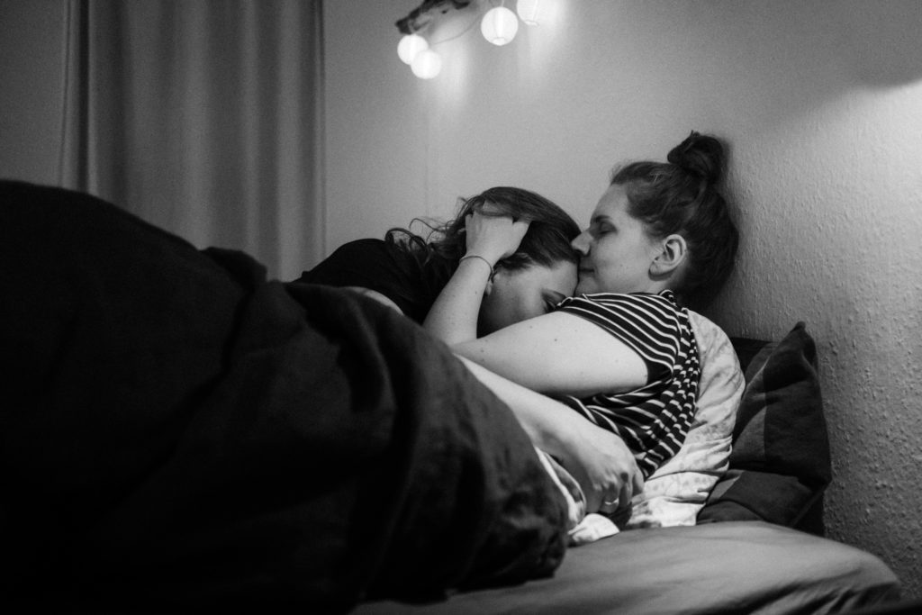 2 Frauen liegen in einem Bett, sie umarmen sich, der Kopf einer Frau liegt auf dem Oberkörper der anderen Frau, beide schließn die Augen