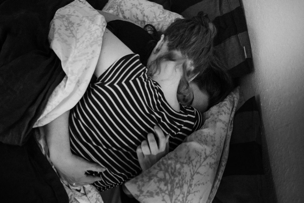 2 Frauen liegen in einem Bett, sie umarmen sich, ihre Köpfe schmiegen sich aneinander